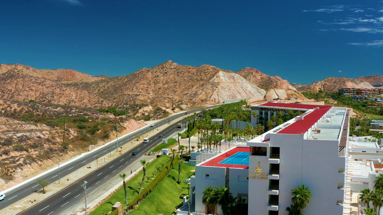 At 0:02, the elevated composition keeps the highway on the left, the white building and palms on the right, and tan mountain ridges across the background, with no visible people.