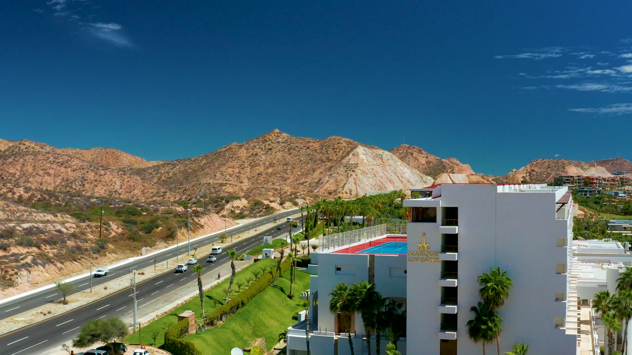 Opening frame at Marquis Los Cabos shows light traffic on a multi-lane road, a white building with rooftop pool area, palm trees, desert mountains, blue sky, and no visible people.