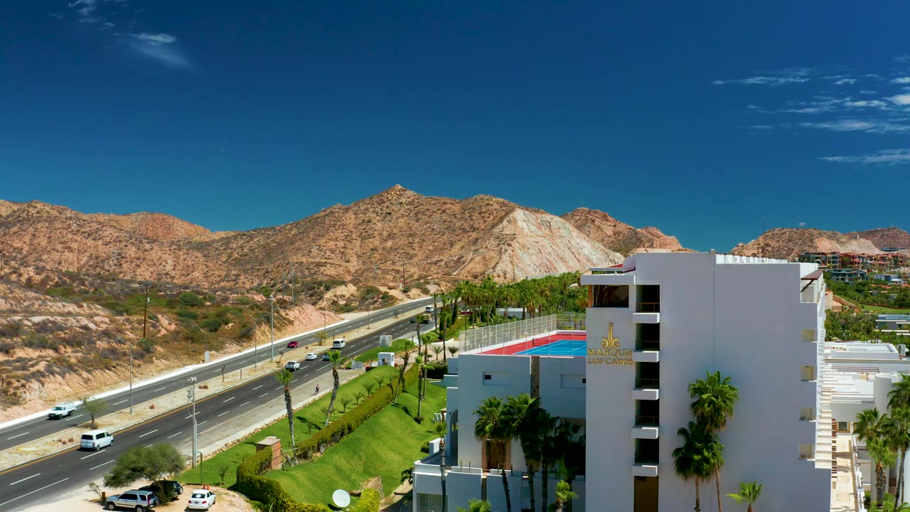 At 0:02, vehicles on the Marquis Los Cabos highway are more spread across the lanes, with palm trees, a rooftop pool, desert slopes, and no visible people in frame.