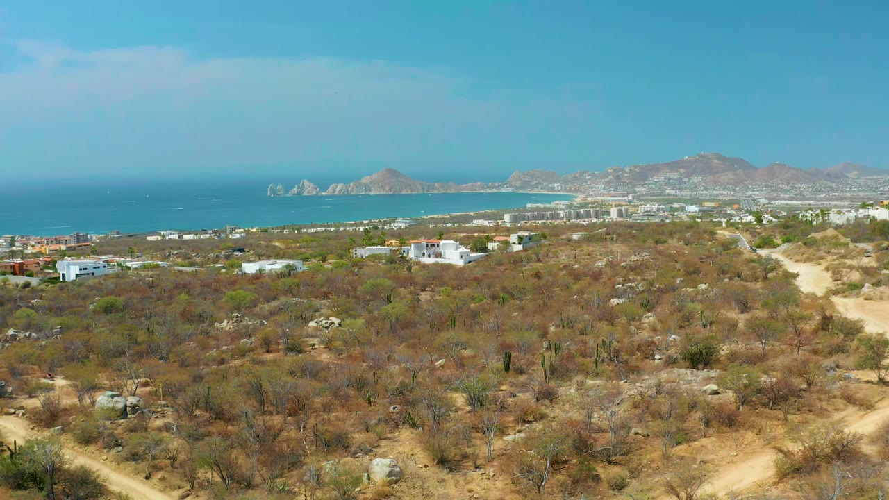 Opening frame of Cabo San Lucas coastal desert with dry scrub and dirt tracks in the foreground, scattered buildings near the shore, blue ocean beyond, and no visible people.