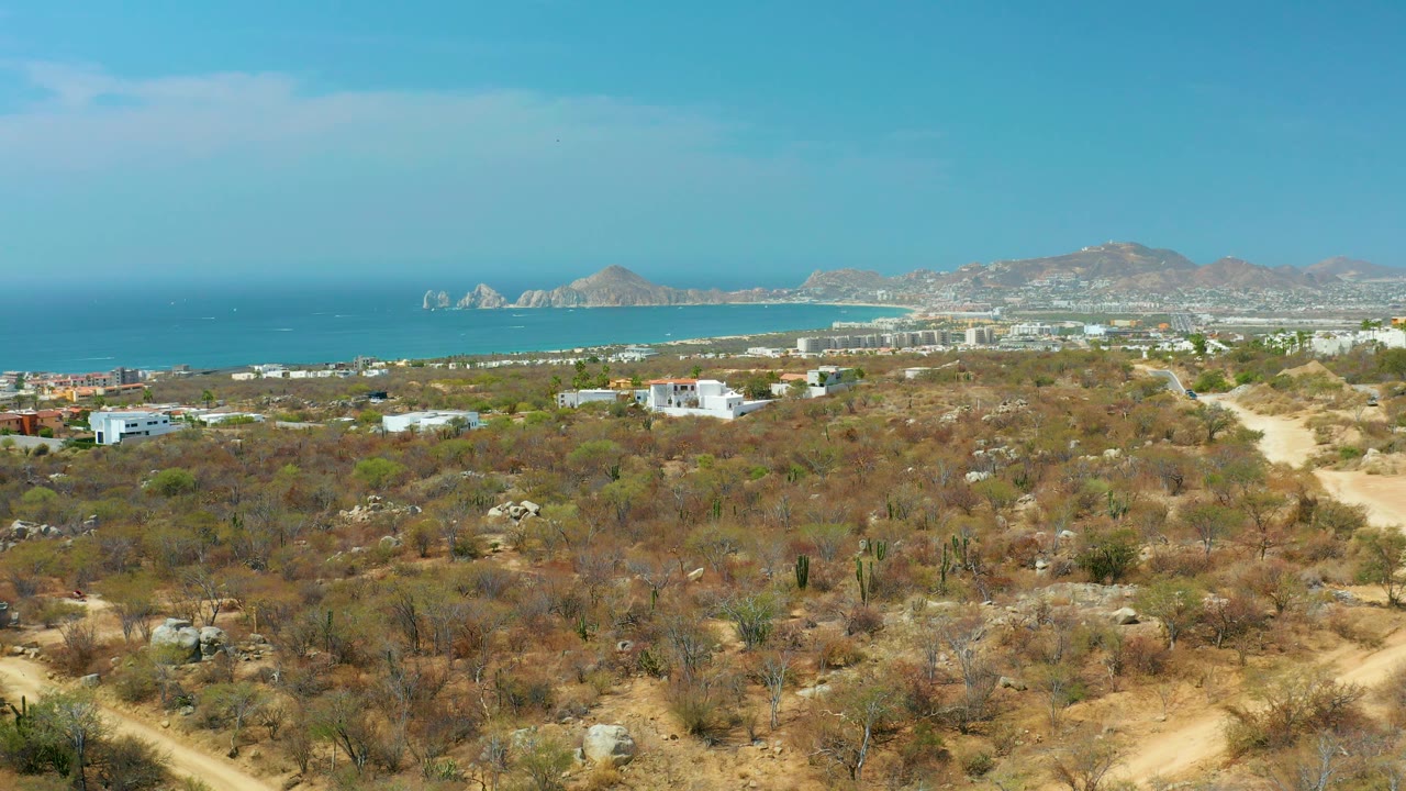 Cabo San Lucas frame showing sparse desert vegetation below, small buildings across the midground, a calm blue ocean band, low mountains on the horizon, and no visible people.