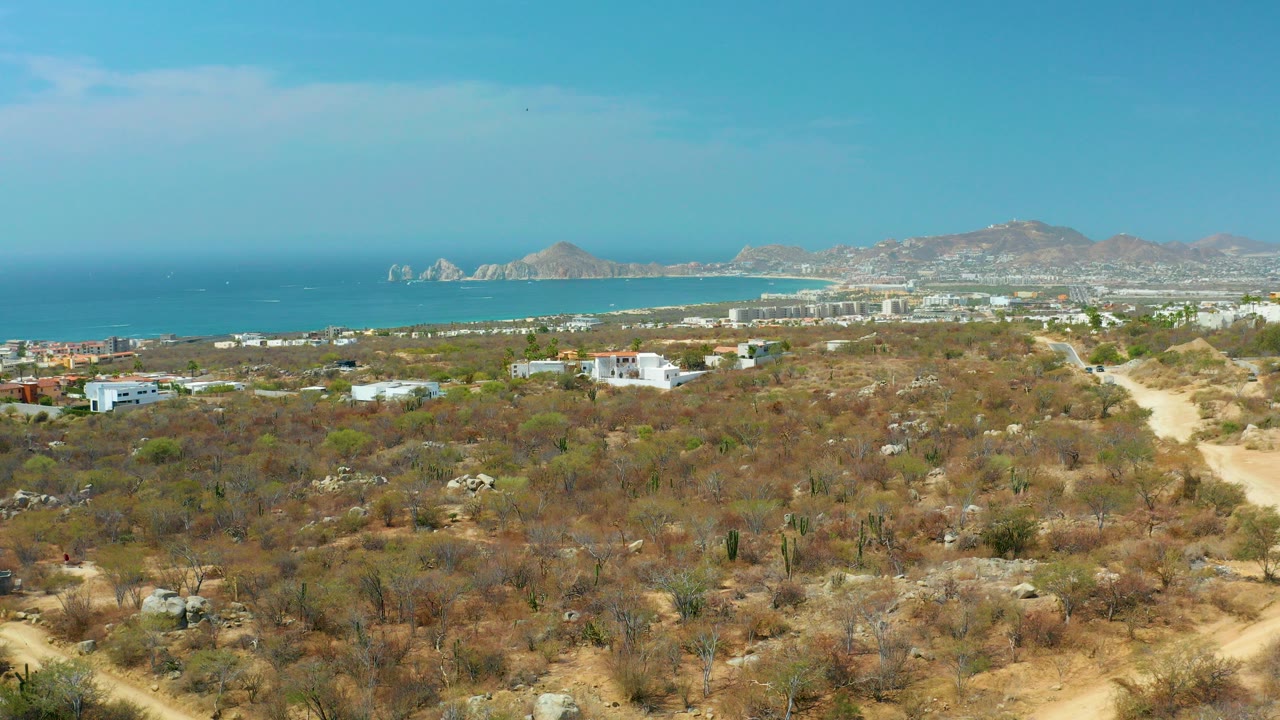 Opening frame of Cabo San Lucas, Baja California Sur, with dry scrubland in front, a pale dirt road on the right, white coastal buildings, blue ocean, distant hills, and no visible people.