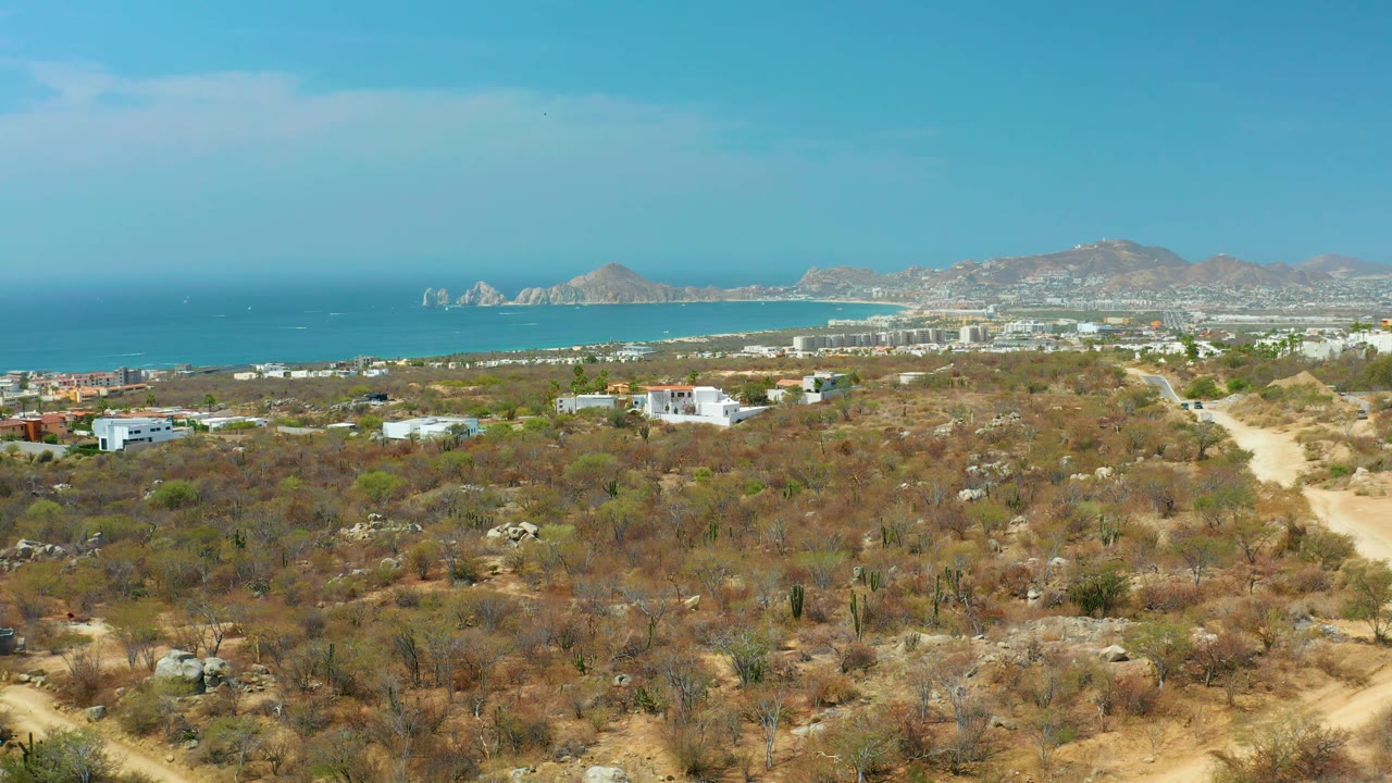 Wide Cabo San Lucas coastal landscape showing dry brush and cactus-like vegetation in the foreground, a road climbing along the right edge, low buildings near the shore, blue ocean, distant hills, and no visible people.