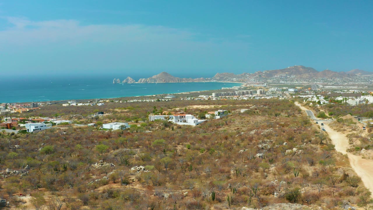 Opening aerial frame over Cabo San Lucas dry scrubland with a pale dirt road, scattered low buildings, blue ocean, rocky headlands, distant mountains, and no visible people.