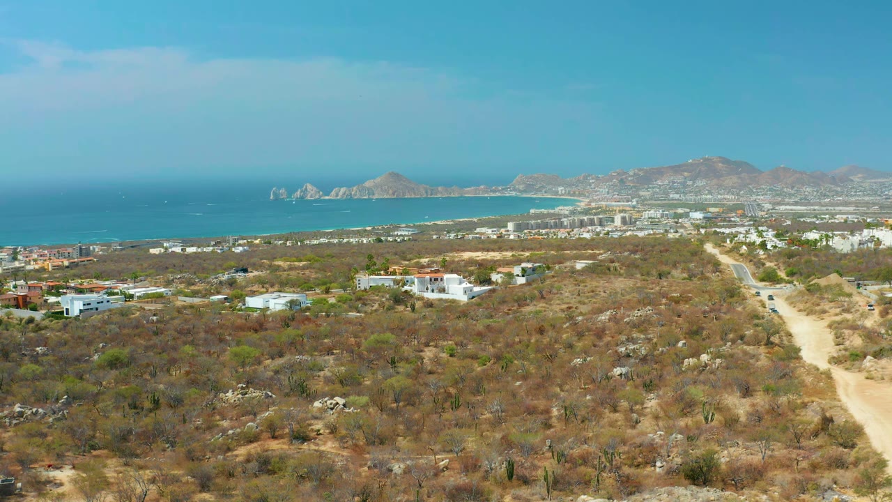 Aerial frame in Cabo San Lucas showing arid foreground brush, a dirt track, white buildings in the midground, bright ocean water, coastline, mountains, and no visible people.