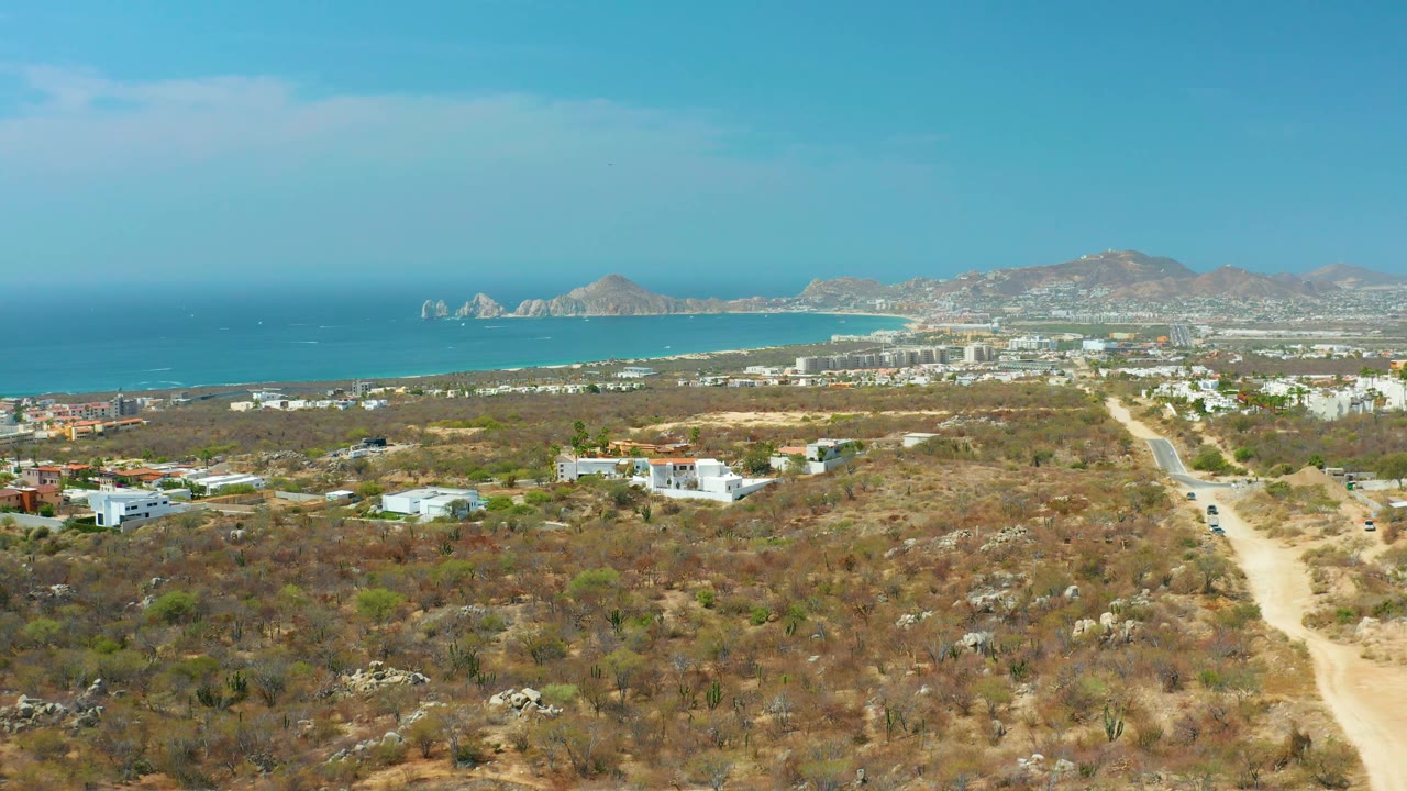 Dry coastal terrain with a dirt road, small buildings, ocean horizon, and mountain backdrop.