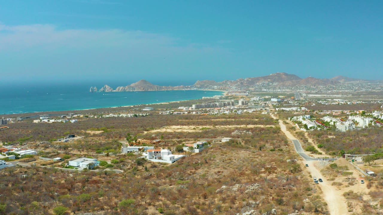 Wide aerial view in Cabo San Lucas, Mexico, with dry foreground terrain, a central road, scattered white houses, blue ocean, distant mountains, and no visible people.