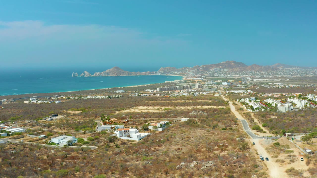 Aerial view across Cabo San Lucas with a pale road through dry land, low white buildings, moving vehicles, ocean water on the left, mountain slopes on the horizon, and no visible people.