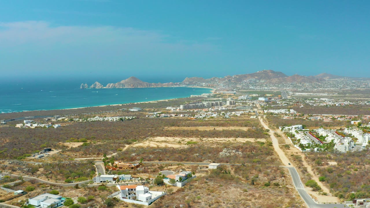 Opening aerial frame over Cabo San Lucas with dry scrubland in front, pale roads and white buildings in the middle distance, blue ocean and mountains beyond, and no visible people.