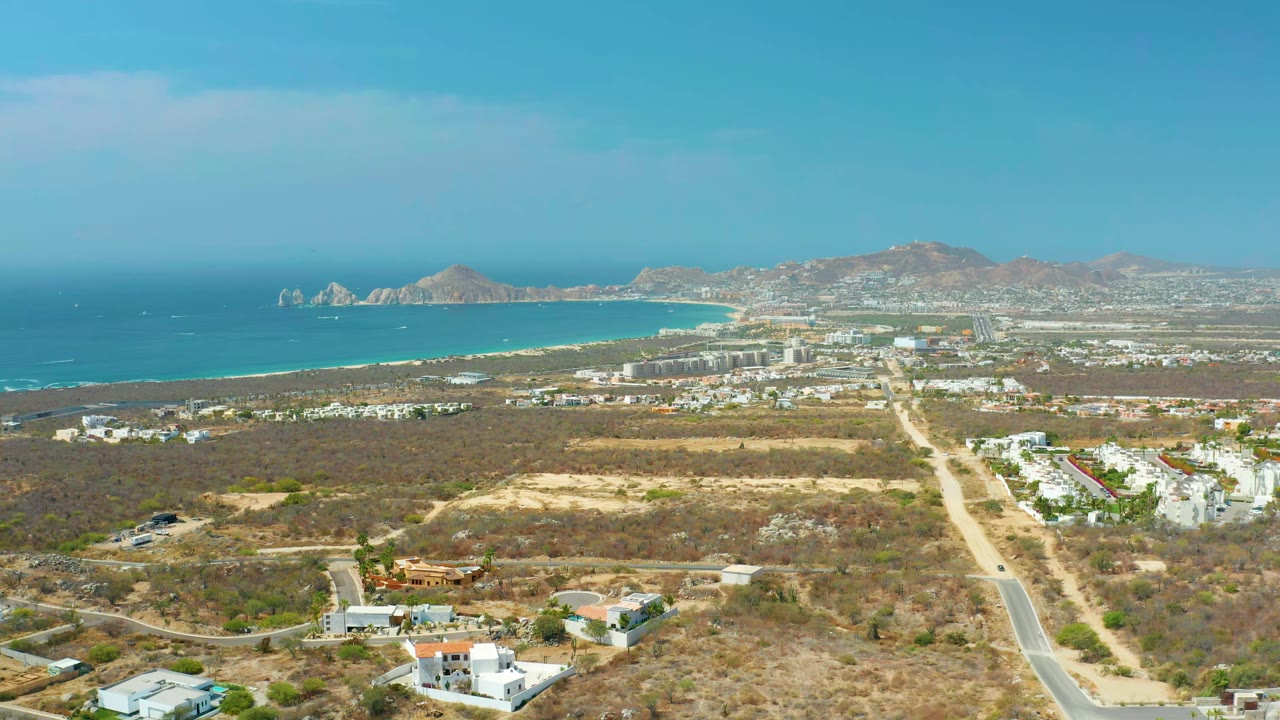 Cabo San Lucas aerial view at 0:01 showing the central road more prominently through dry terrain, with coastal buildings, turquoise water, mountains, and no visible people.