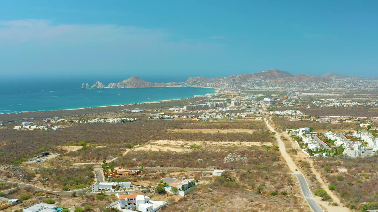 Cabo San Lucas frame at 0:02 with dry lots and roads in the foreground, low white buildings to the right, a curving beach, blue ocean, mountains, and no visible people.