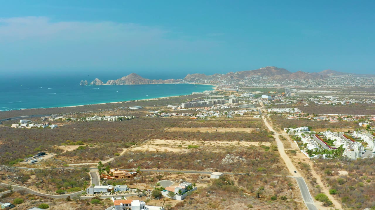 Drone view over scrubland toward coastal buildings, ocean, beach, and distant mountains.