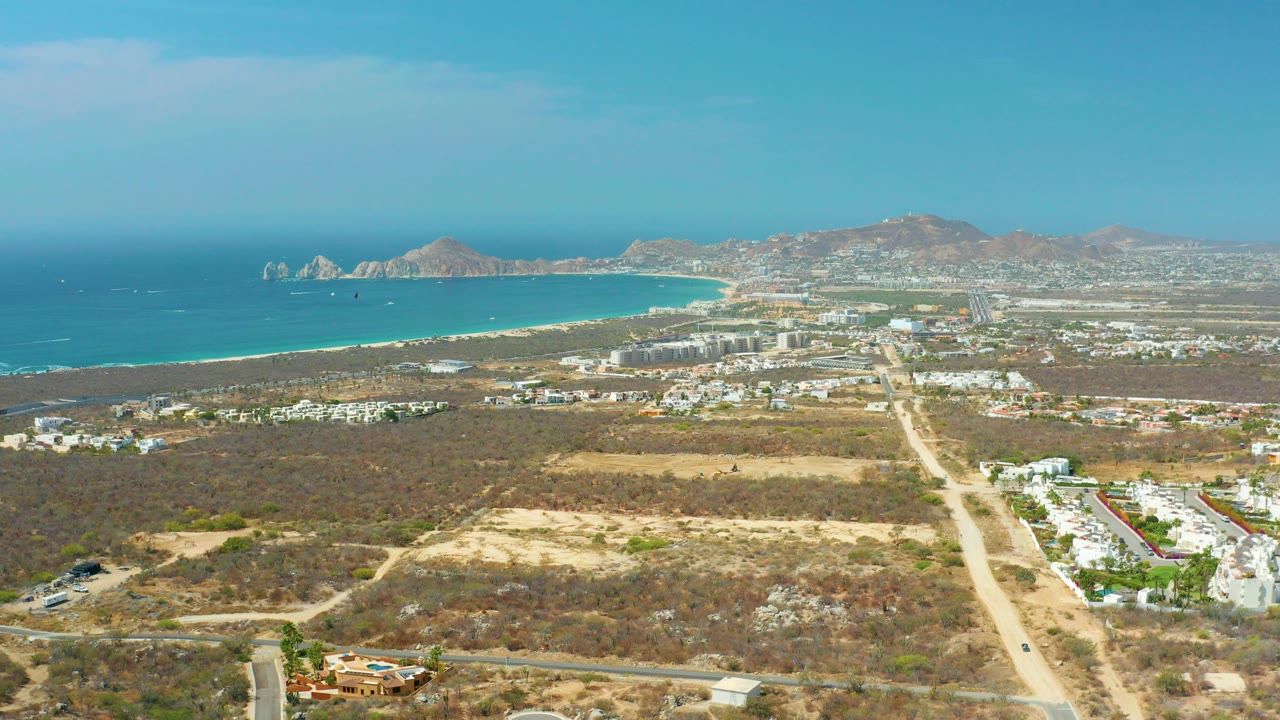 Wide aerial frame over Cabo San Lucas with dry foreground terrain, a pale road leading toward white buildings, turquoise ocean, sandy beach, distant mountains, and no visible people.