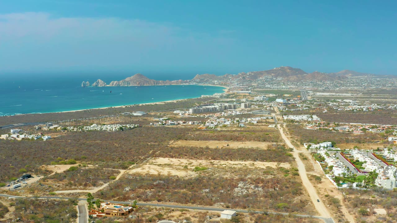 Aerial view in Cabo San Lucas showing the coastline stretching left to right, white buildings near the beach, dry scrubland in the foreground, and no visible people.