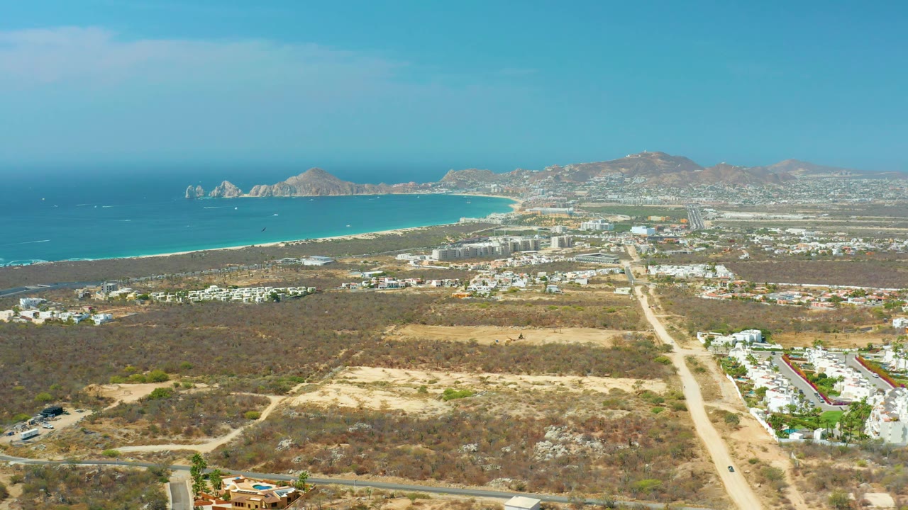 High drone-style frame over Cabo San Lucas with a bright blue ocean, pale beach, low mountain horizon, scattered buildings, a central dirt road, and no visible people.