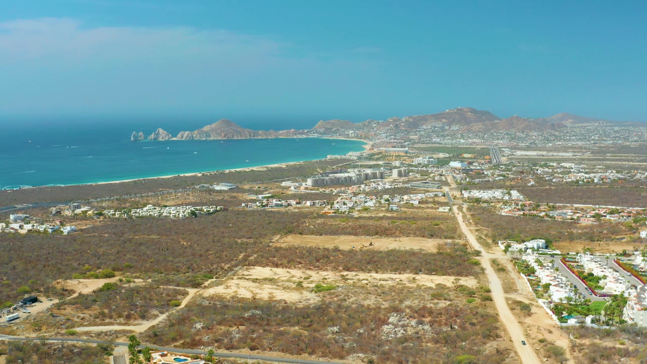 Coastal aerial with arid land, road network, buildings, beach, and ocean.