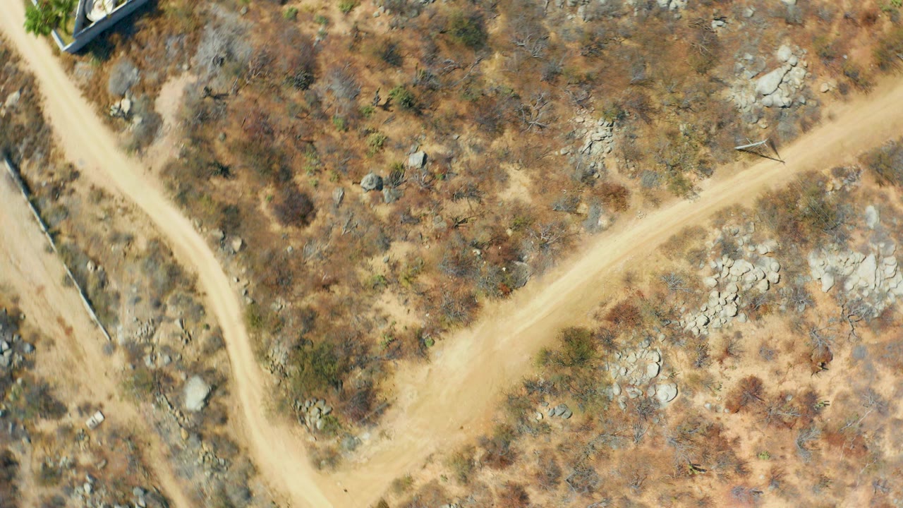 Overhead view in Cabo San Lucas of pale dirt roads intersecting across dry scrub and rocks, with building edges at the corners and no visible people.