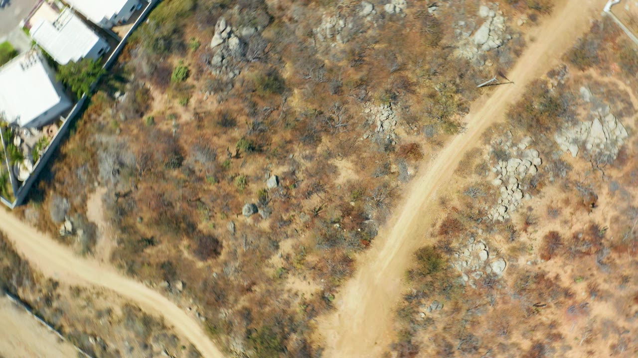Aerial view in Cabo San Lucas showing a curving dirt road through dry vegetation and rocks, with white structures and boundary walls along the upper edge and no visible people.