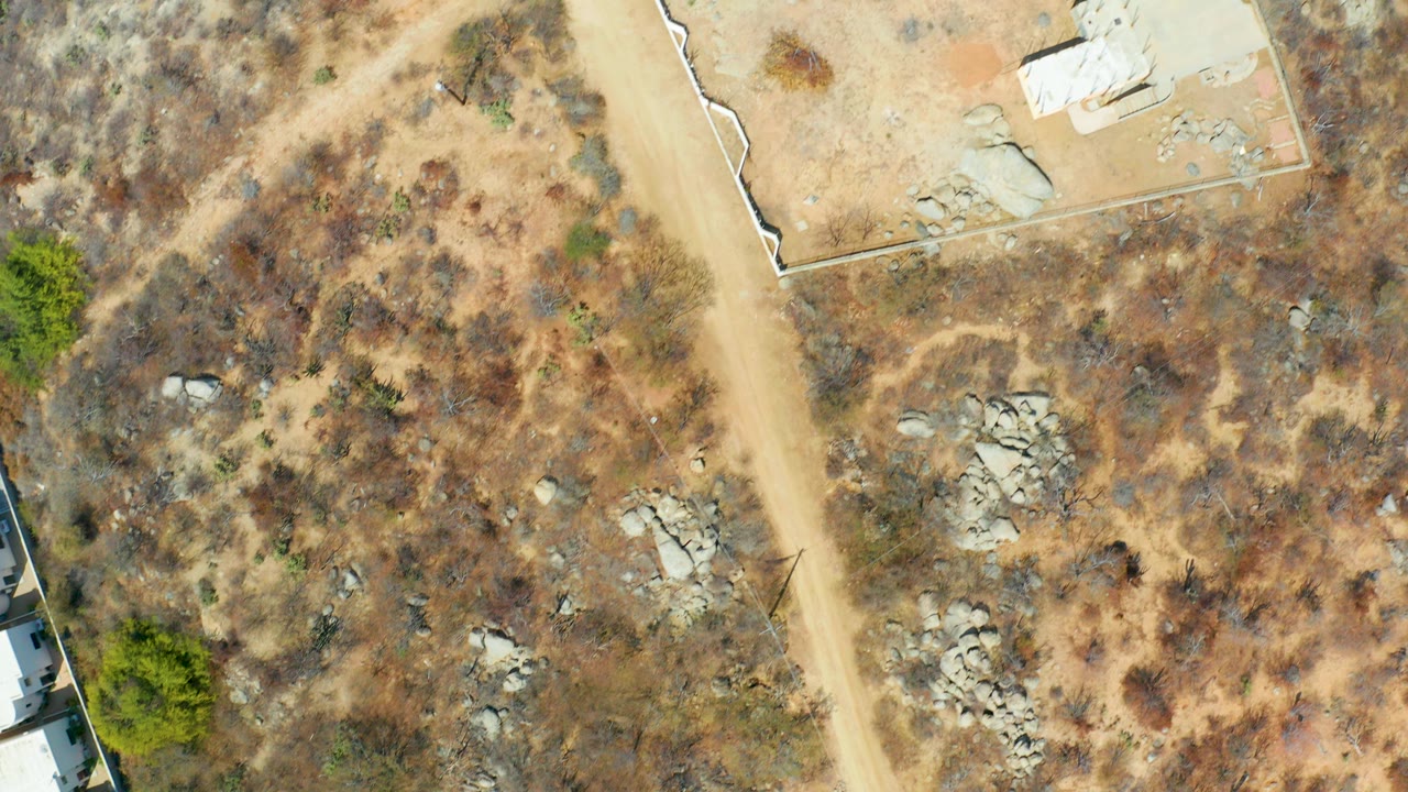 Overhead Cabo San Lucas frame showing a tan dirt road crossing dry scrubland, scattered rock clusters, a partial pale structure at the lower left edge, and no visible people.