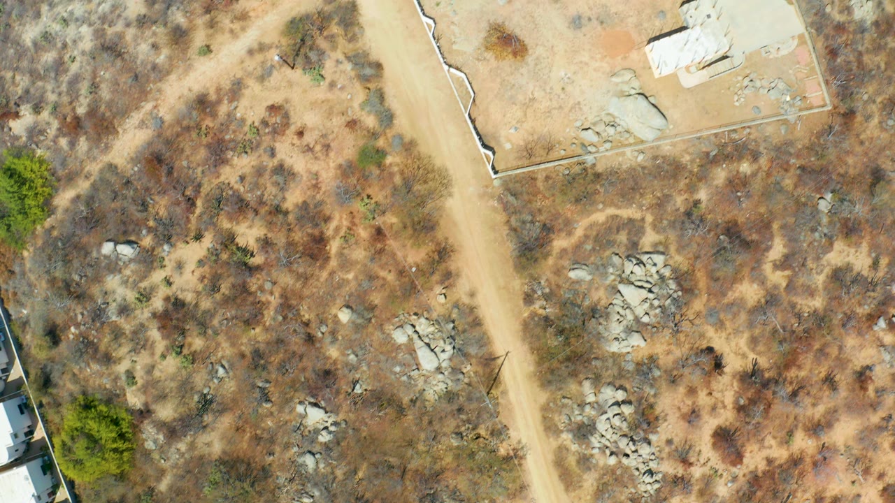 Top-down aerial of a dirt road, rocky scrubland, and a small pale building near the upper edge.