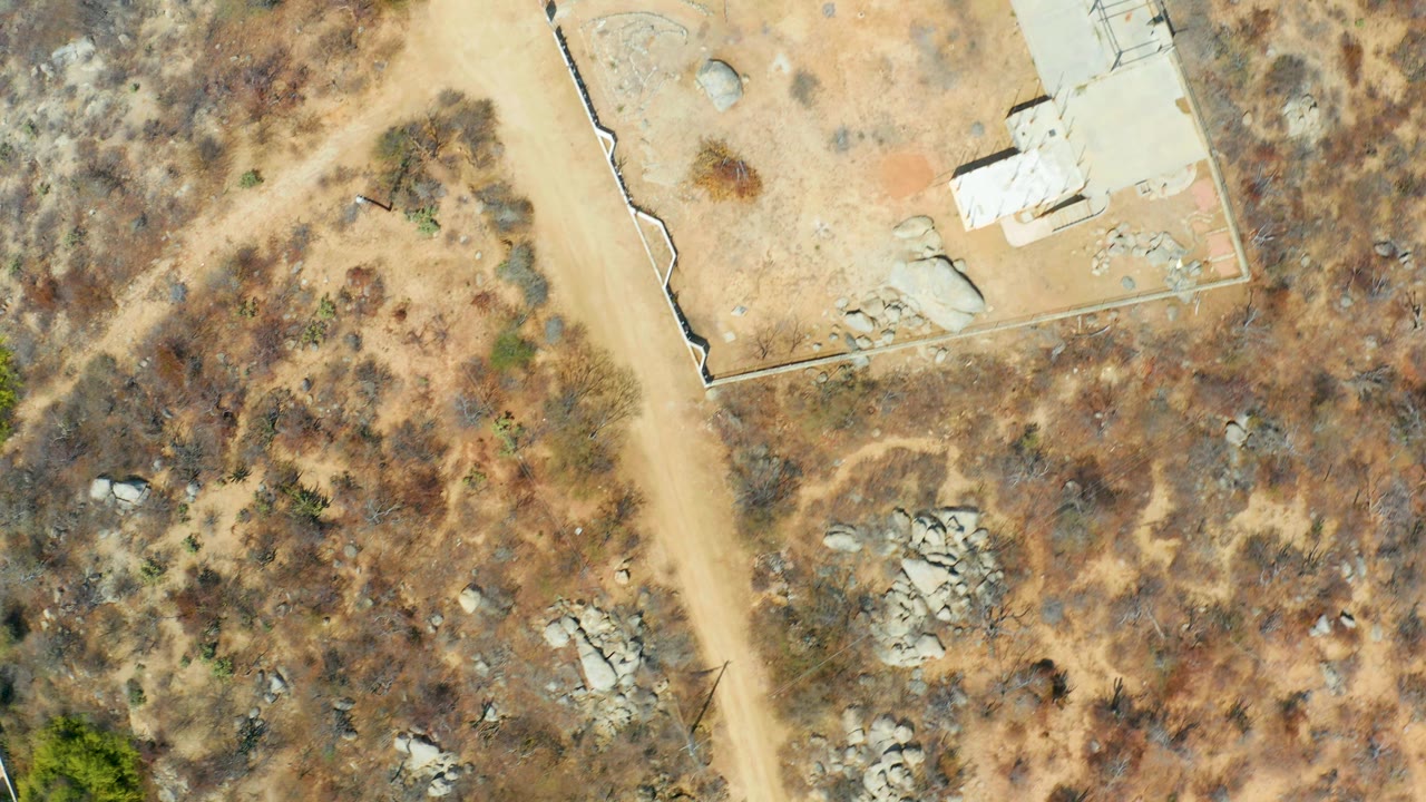 Aerial Cabo San Lucas frame showing dry scrub terrain, a tan road running diagonally, scattered boulders, a pale flat-roof building at the upper right, and no visible people.