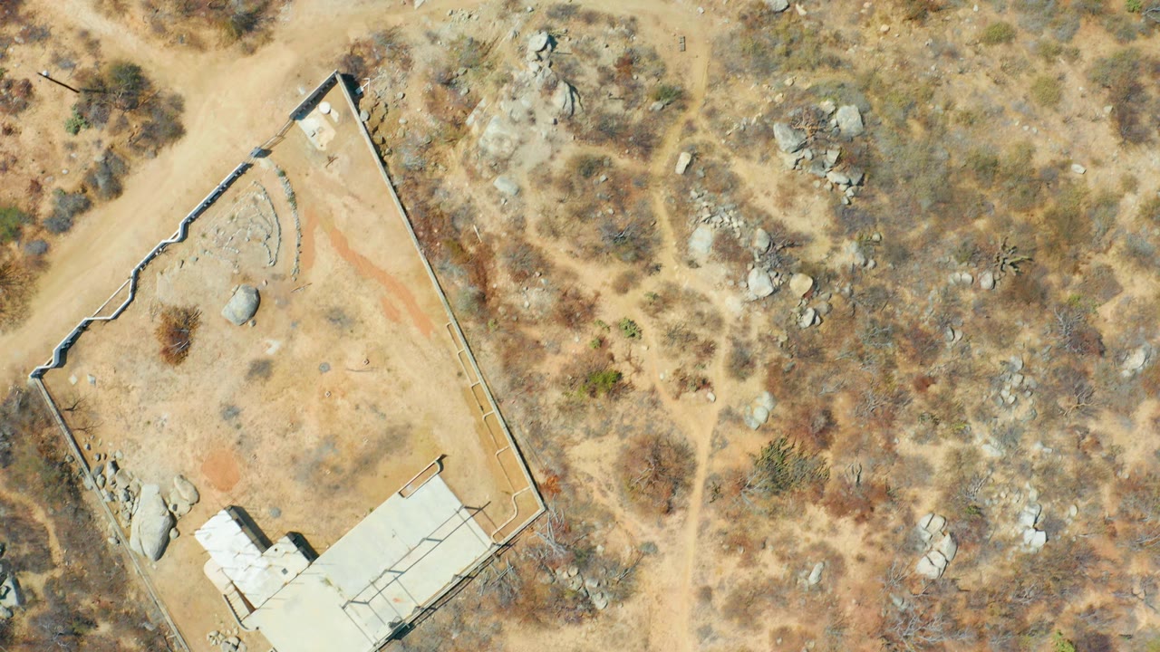 Top-down aerial frame in Cabo San Lucas showing a walled sandy lot, dirt paths, rocks, dry brush, and no visible people.