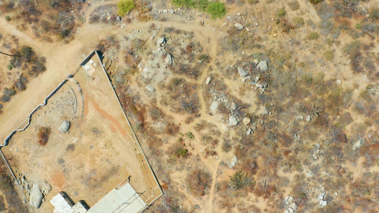 Overhead Cabo San Lucas frame with a tan dirt road curving past a walled property, rocky brushland, small green shrubs, and no visible people.