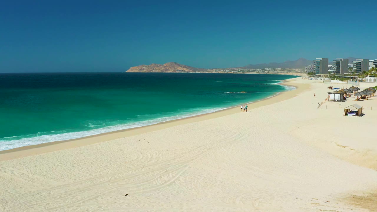 Wide elevated view at Dreams Los Cabos, Baja California Sur, with pale sand in the foreground, turquoise ocean on the left, small walkers near the shore, and resort towers in the distance.