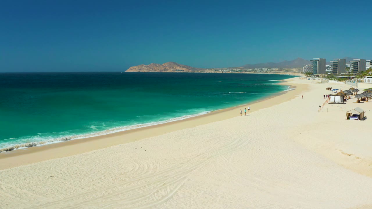 Curving sandy beach beside turquoise ocean with small walkers and buildings in the distance.