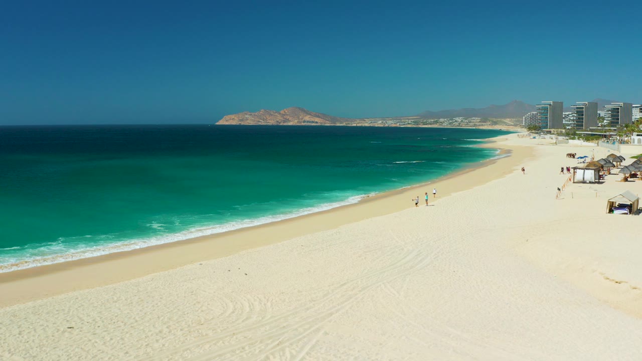 At Dreams Los Cabos, the frame shows more of the right-side beach area with huts or umbrellas, distant hotel buildings, small walkers, and layered turquoise water under a clear sky.