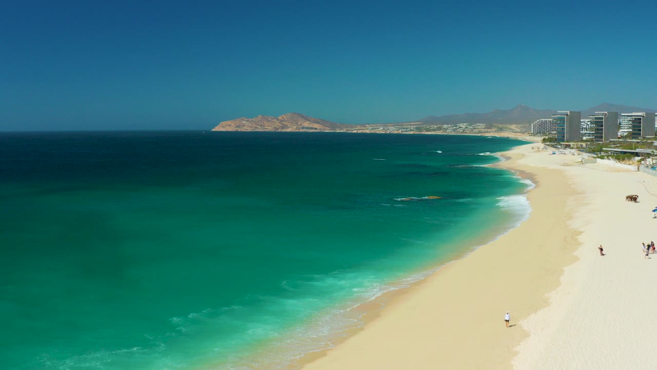 Aerial view at Dreams Los Cabos, Baja California Sur, with turquoise water on the left, a pale sandy beach on the right, distant resort buildings, and tiny people visible near the shoreline.