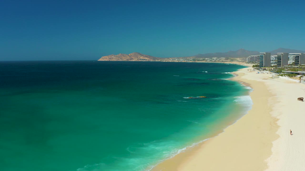 The Dreams Los Cabos shoreline is seen from above with a small rocky patch in the water, pale sand to the right, resort buildings near the edge of frame, and tiny people visible on the beach.