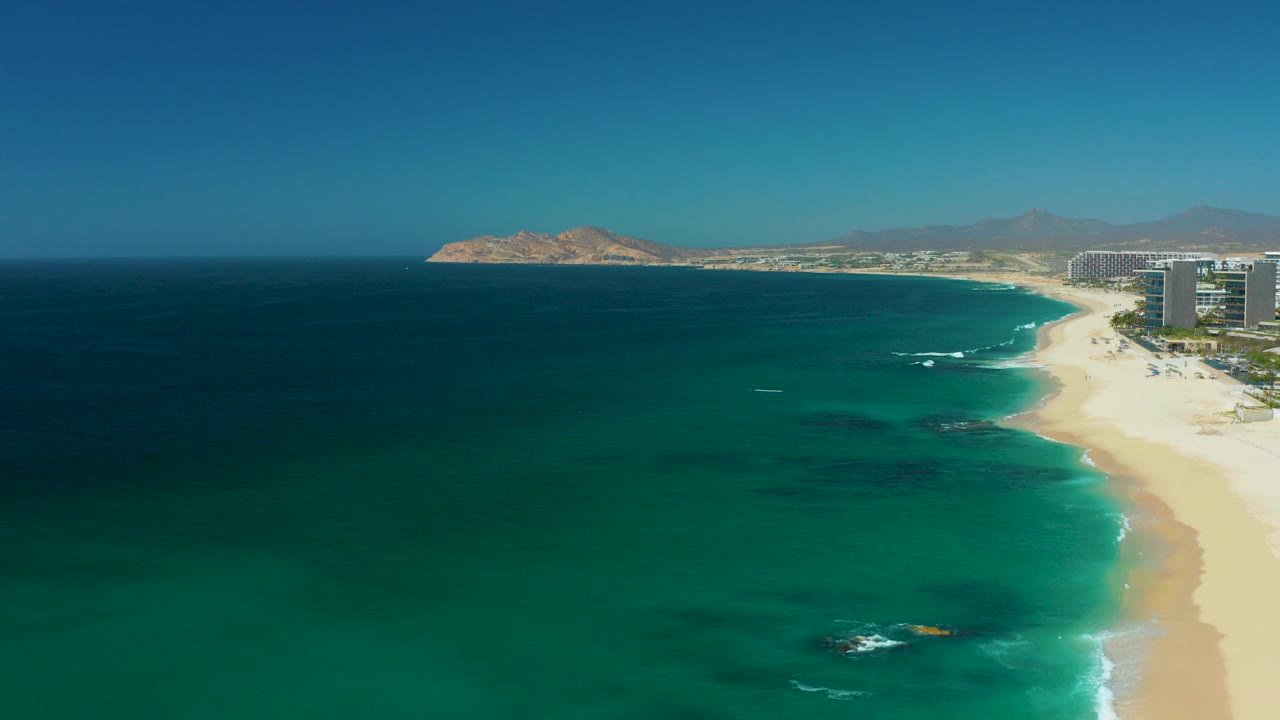 Opening aerial frame at Dreams Los Cabos, Baja California Sur, Mexico, showing turquoise ocean, a pale sandy beach, resort buildings on the right, distant mountains, and no visible people.