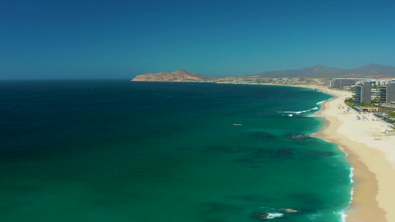 At 0:02, the aerial view over Dreams Los Cabos shows darker reef or rock patches under the turquoise water, a clean sand edge, resort buildings to the right, and no visible people.