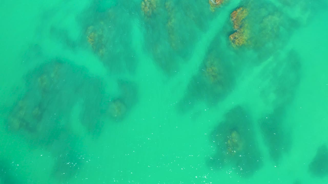 A later overhead frame shows scattered shallow rock formations under turquoise water at Dreams Los Cabos, with fine white particles and no visible people.