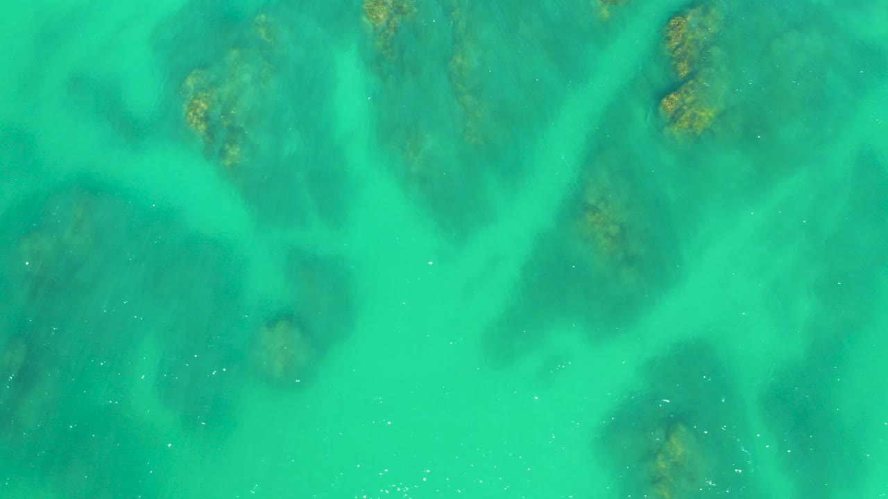 Overhead view of turquoise shallow ocean at Dreams Los Cabos with soft ripples, dark underwater patches, and no visible people.