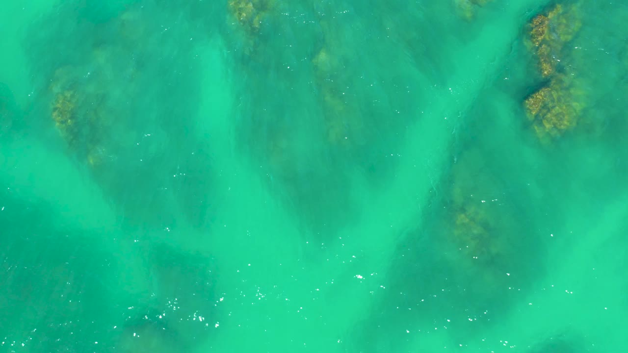 Aerial frame over Dreams Los Cabos water with a darker submerged rock mass toward the lower left, soft green-blue water bands, scattered white specks, and no visible people.