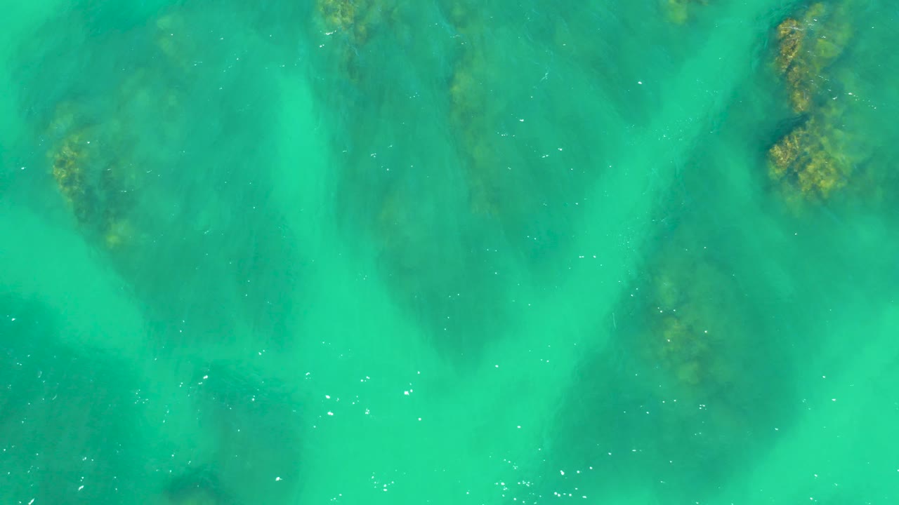 Top-down Dreams Los Cabos water view with submerged rocks clustered near the upper edge, darker green underwater areas, fine surface ripples, and no visible people.