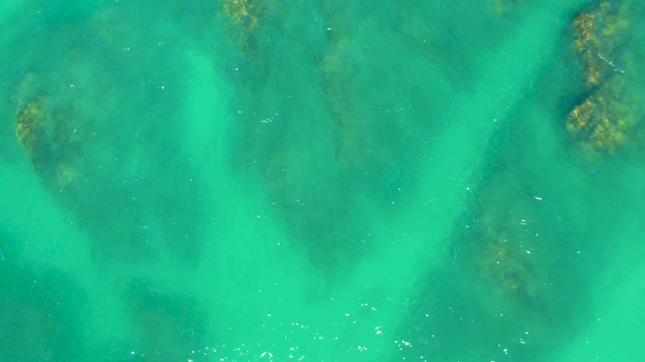 Straight-down frame from Dreams Los Cabos showing shallow turquoise water, diffuse dark patches below the surface, faint foam marks, and no visible people.