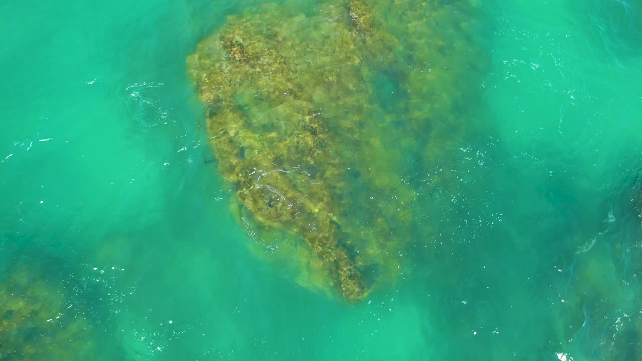 The rock sits high in the frame under clear turquoise seawater at Dreams Los Cabos, with faint white ripples crossing the surface and no visible people.