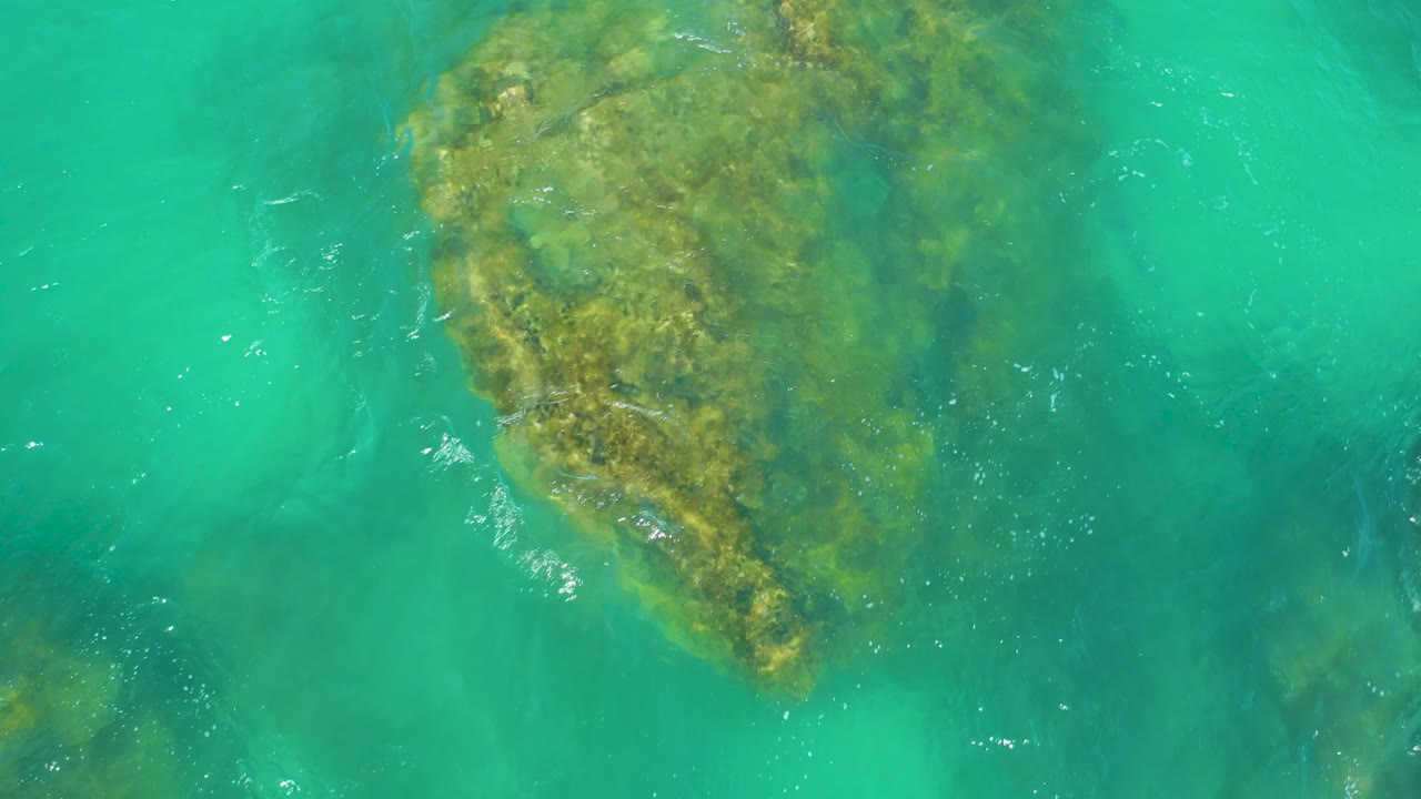 A large algae-stained rock remains visible beneath teal-turquoise water at Dreams Los Cabos, with brighter ripples along one edge and no visible people.