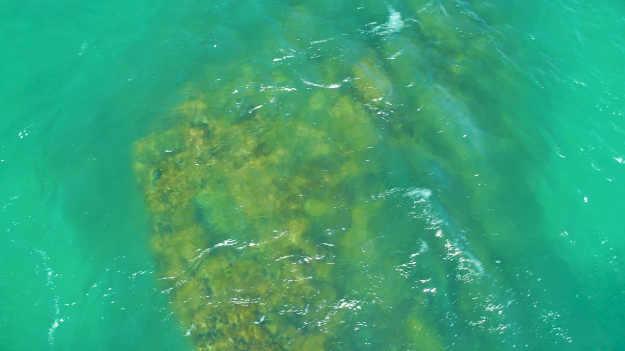 Overhead view of turquoise water over yellow-green submerged rocks at Dreams Los Cabos, with ripples on the surface and no visible people.