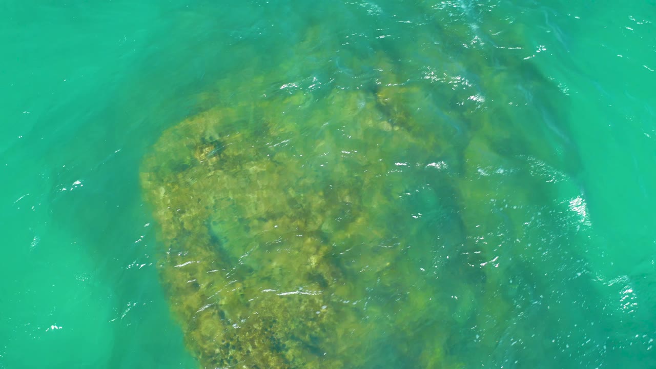 A submerged yellow-green rock edge sits under clear turquoise water at Dreams Los Cabos, with faint foam marks and no visible people.