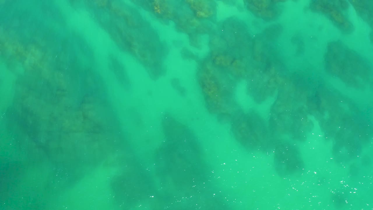Straight-down aerial view of turquoise ocean water with darker seabed formations and small surface glints at Dreams Los Cabos, with no visible people.