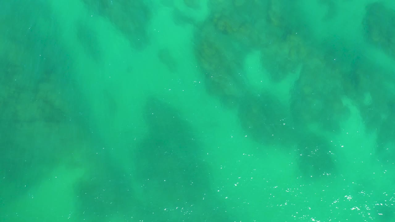 Overhead shallow ocean water at Dreams Los Cabos shows darker rock forms beneath brighter turquoise water as ripples begin to blur the bottom detail, with no visible people.