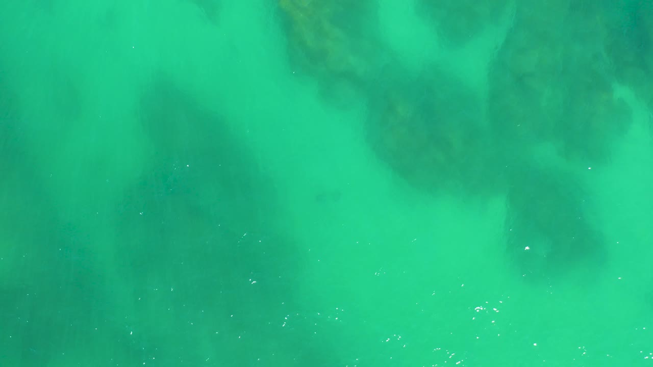 Turquoise ocean water covers soft-edged dark seabed shapes at Dreams Los Cabos in Mexico, with tiny bright reflections and no visible people.