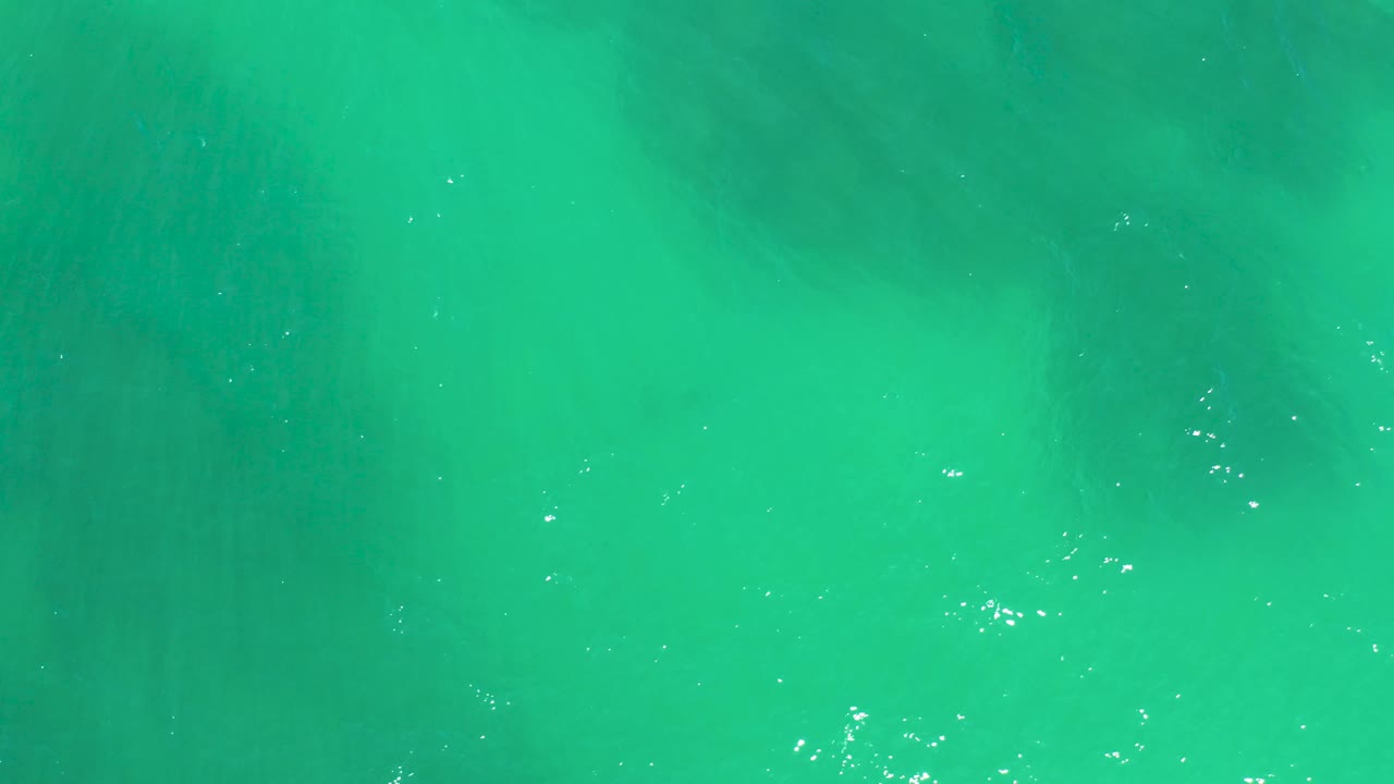 A top-down frame at Dreams Los Cabos shows turquoise ocean surface over darker submerged rock areas, with thin ripples and no visible people.