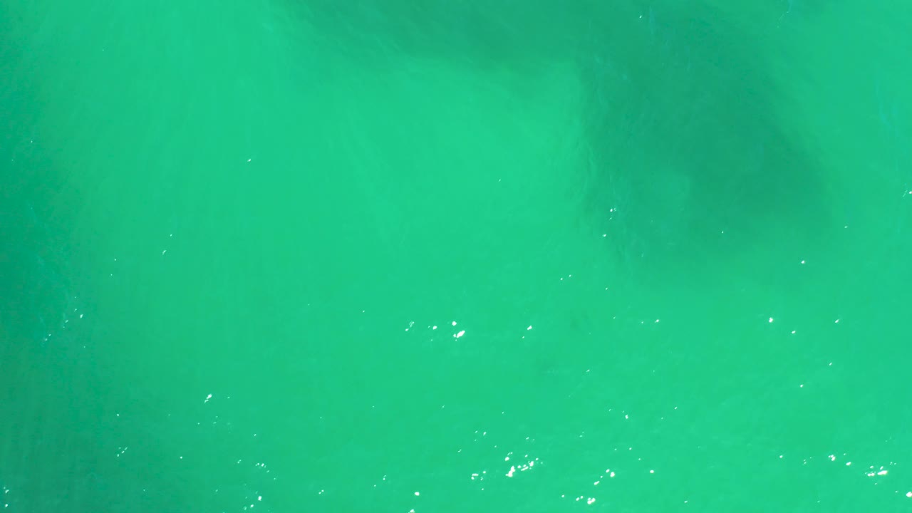 At 0:02, the overhead view shows clear turquoise water at Dreams Los Cabos, scattered bright reflection points, soft underwater tonal bands, and no visible people.