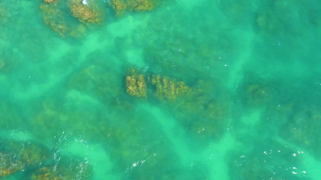 Clear green-blue ocean water reveals darker submerged rock patches at Dreams Los Cabos, with surface shimmer and no visible people.