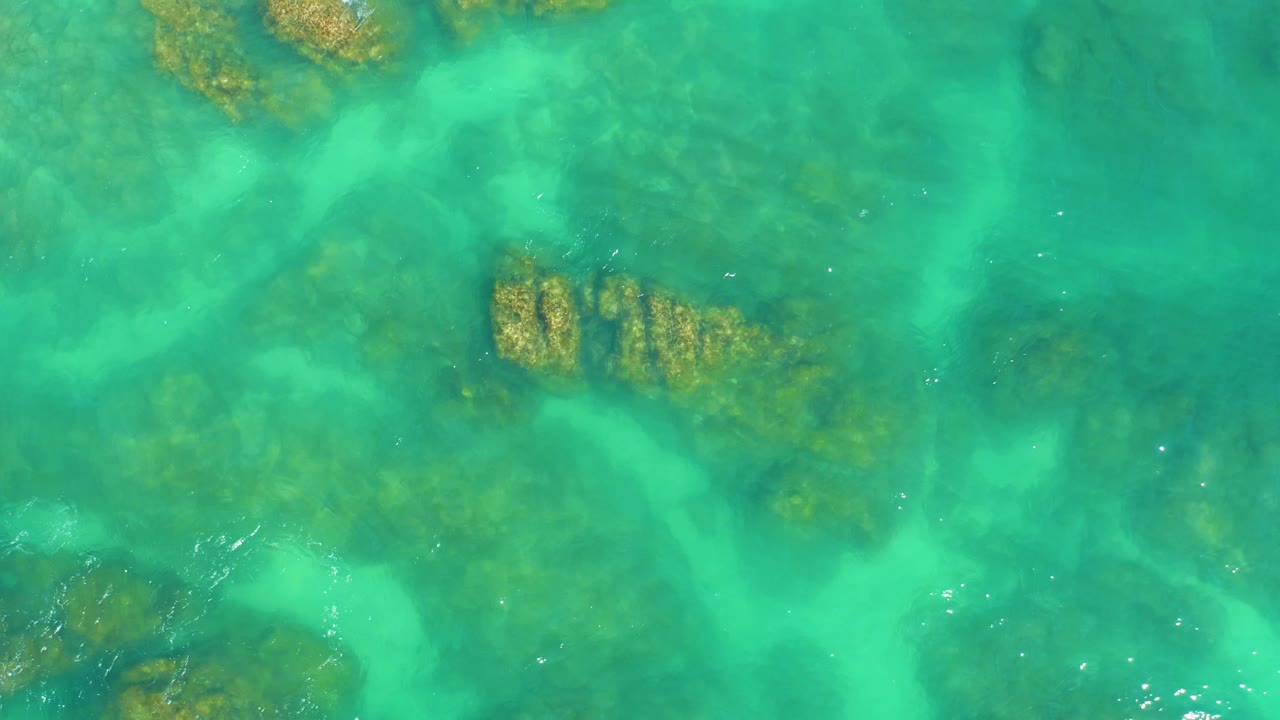 Rippled turquoise water covers a rocky seabed at Dreams Los Cabos, with a larger rock cluster entering the left side and no visible people.